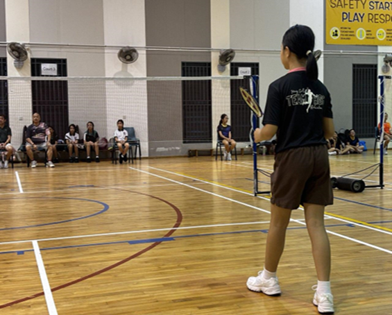 A young girl holding a badminton racket stands on an indoor court, facing the net, with spectators seated along the wall.