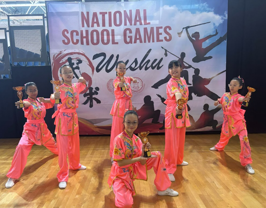 Six girls in pink wushu uniforms pose with trophies in front of a National School Games Wushu banner.