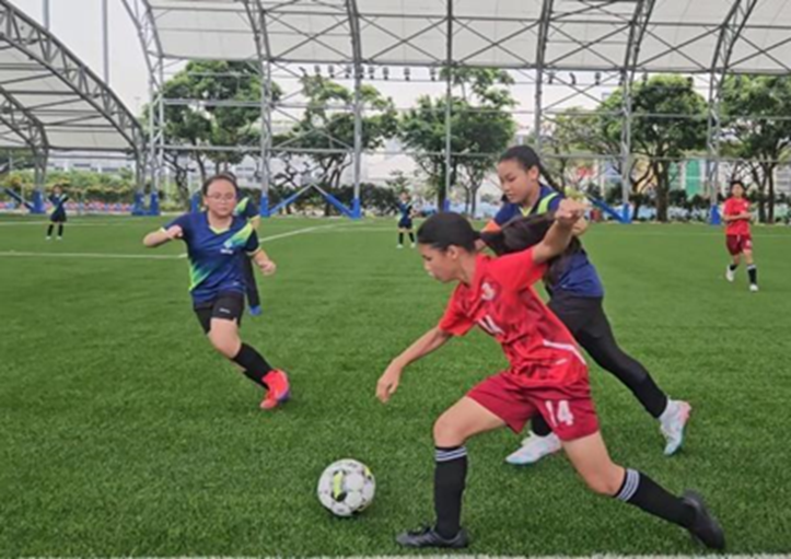 Girls in red and blue uniforms compete for a soccer ball on a green turf field under a large canopy structure.