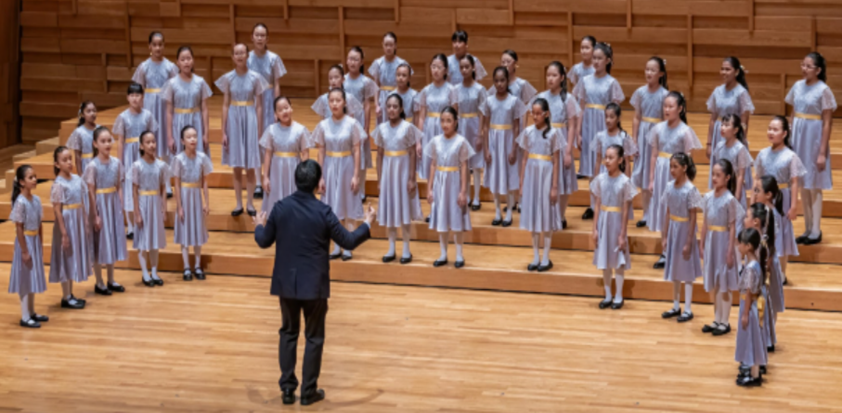 A conductor in black leads a large girls choir in matching silver dresses with gold sashes on a wooden stage.