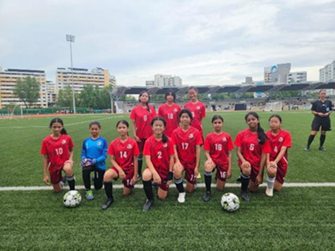 Girls soccer team in red jerseys posing on a green field with two soccer balls, a referee stands nearby.