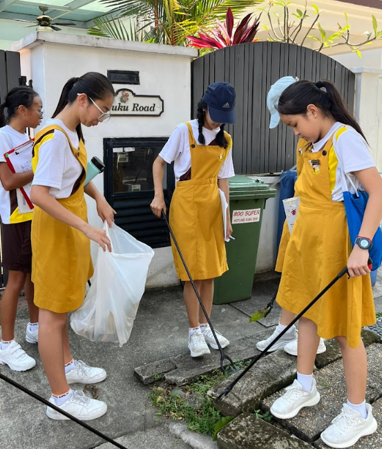 Three primary school girls in yellow pinafore uniforms conduct a litter-picking activity along a residential street. Two girls use long-handled litter pickers to collect rubbish from the ground, while a third holds open a clear plastic bag to collect the waste. Green rubbish bins and a road sign reading "Tuku Road" are visible in the background.