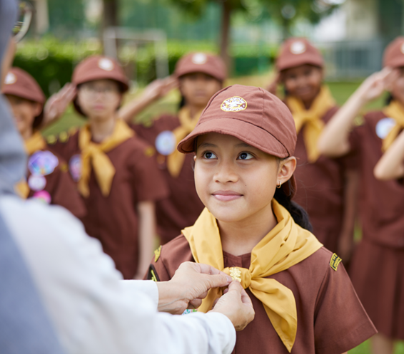 A girl in a brown scout uniform receives a badge while others salute in the background outdoors.