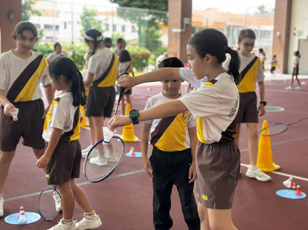 Students in brown and yellow uniforms practice badminton on an outdoor court with orange cones and shuttlecocks.