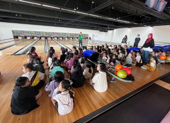 A large group of children sitting on the floor of a bowling alley, listening to an instructor standing at the far end of the lanes. Several adults and chaperones stand nearby.