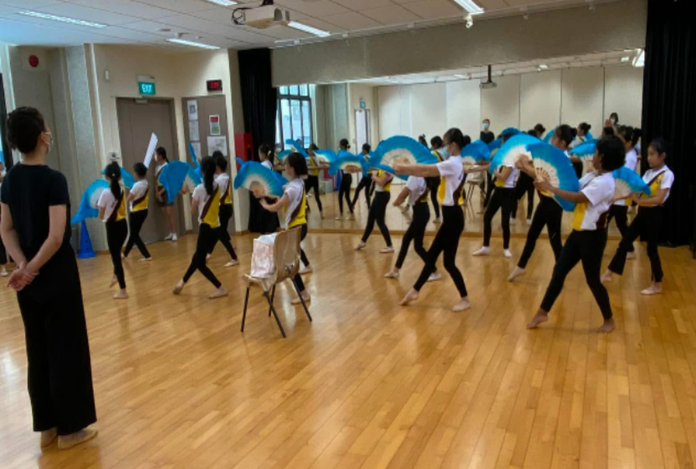 Girls in white and black outfits practice a fan dance with large blue fans in a mirrored studio.