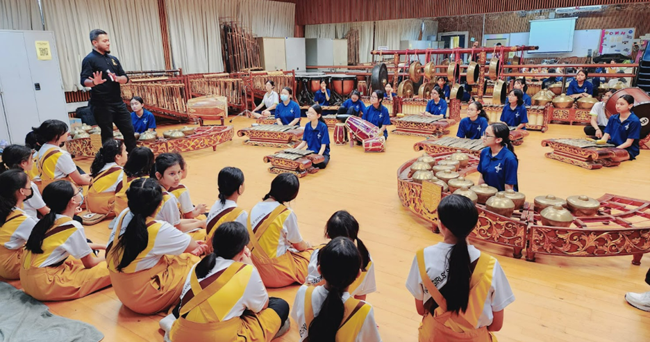 Students in yellow vests sit on the floor watching a gamelan music lesson with traditional Indonesian instruments.