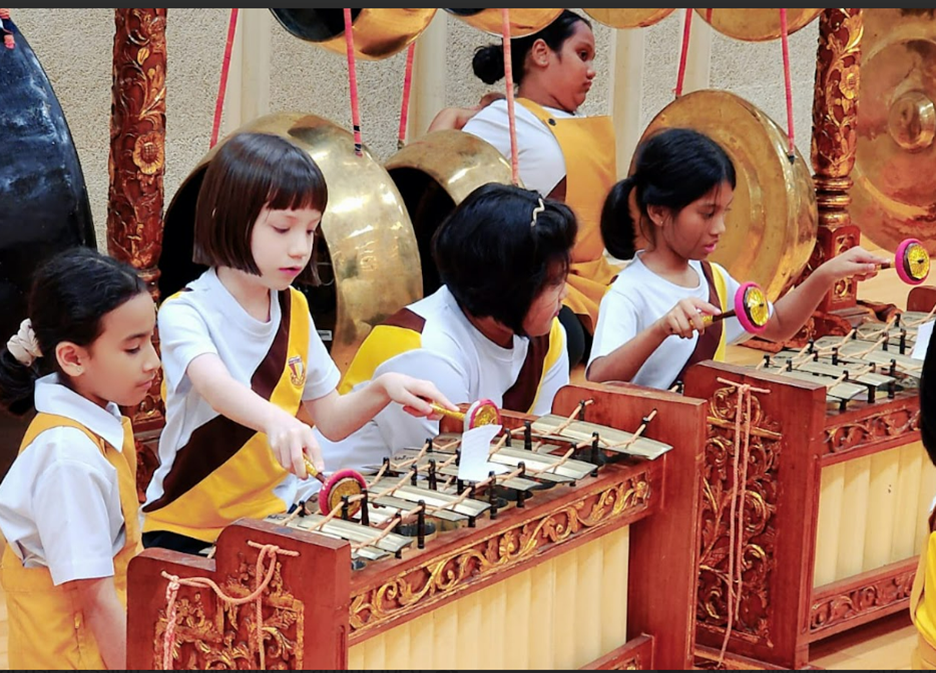 Children in yellow and white uniforms playing gamelan instruments with large brass gongs hanging in the background.