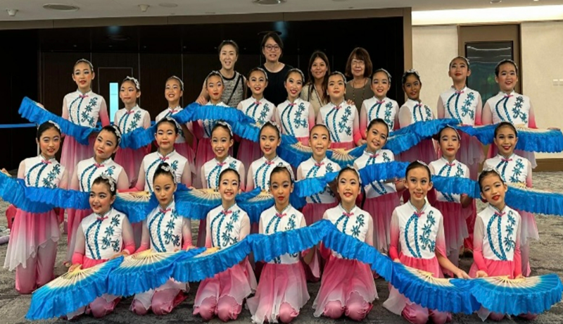 A large group of young female dancers in white and pink costumes holding blue decorative fans pose together.