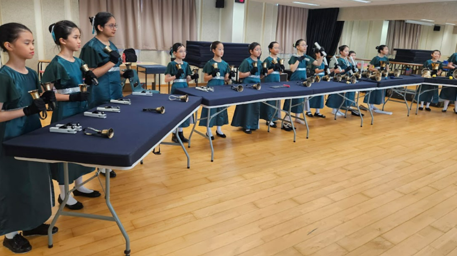 Girls in teal dresses wearing black gloves stand along tables holding handbells in a rehearsal room.
