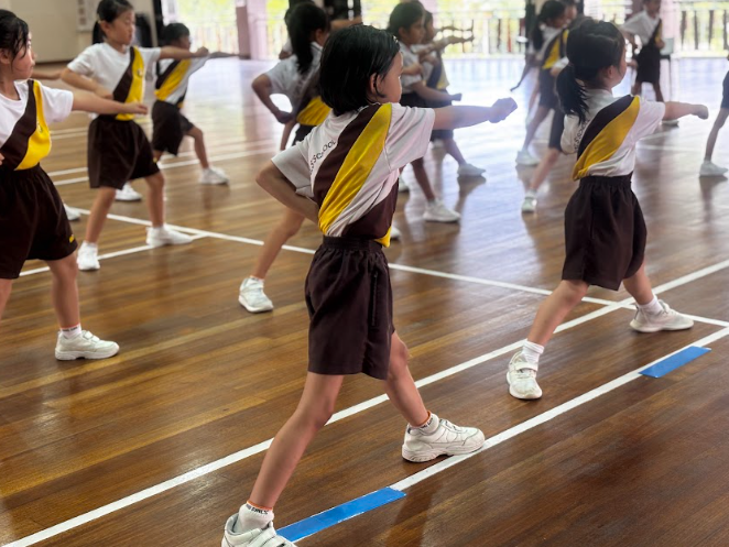 A larger group of girls in the same school PE uniforms performing a synchronised martial arts or dance routine in a school hall, viewed from behind.