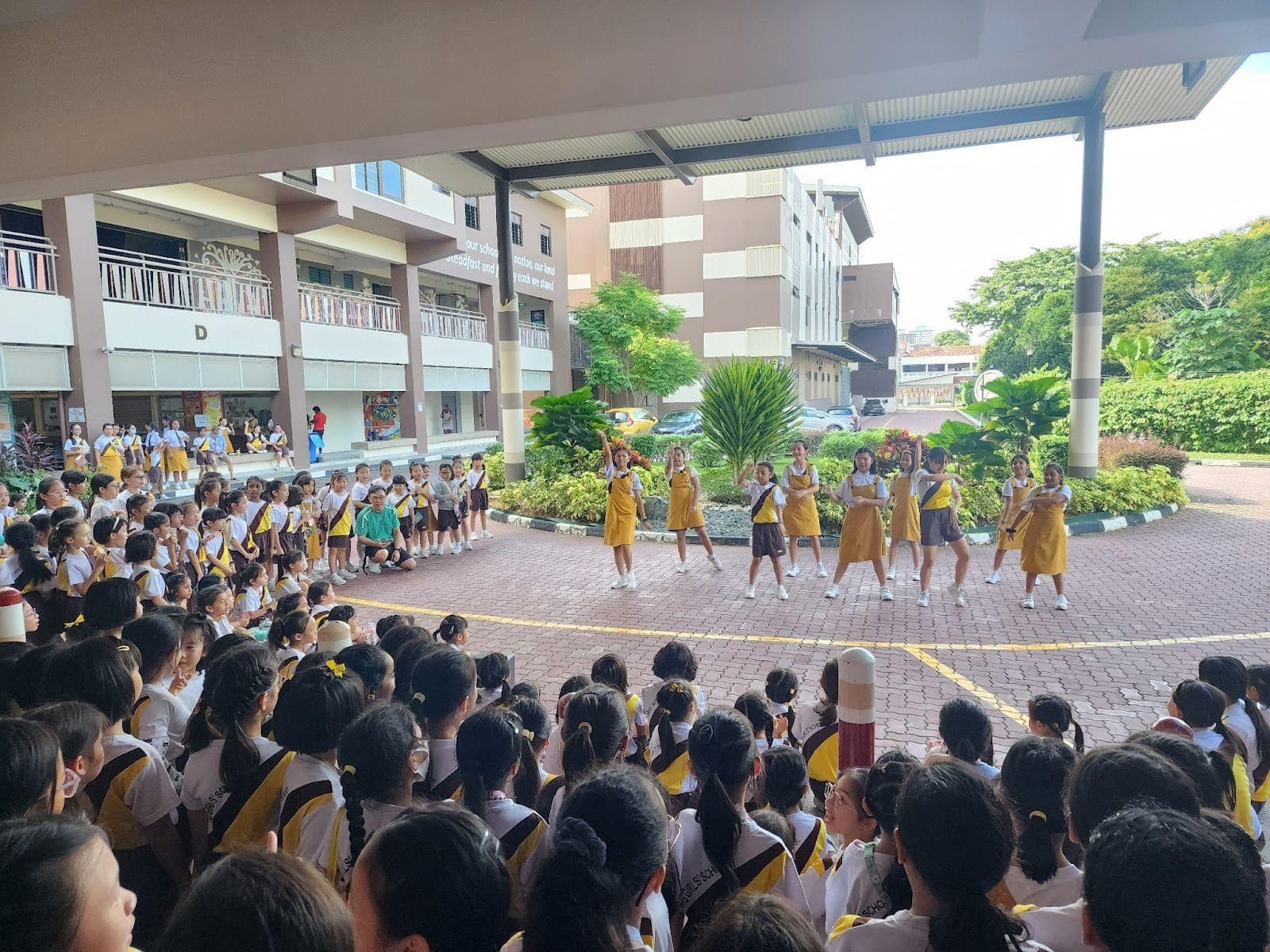 A large group of primary school girls in white and yellow-brown uniforms gather in an open-air school courtyard to watch a group of older students perform a dance routine.