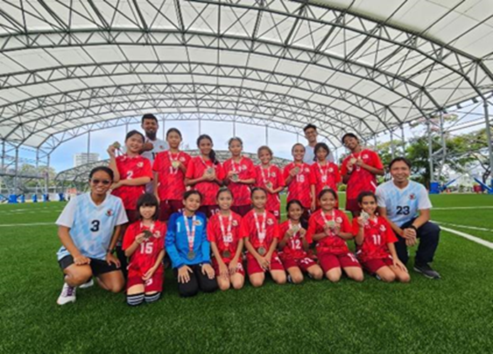 A youth girls soccer team in red uniforms poses with medals on a green turf field under a large canopy structure.