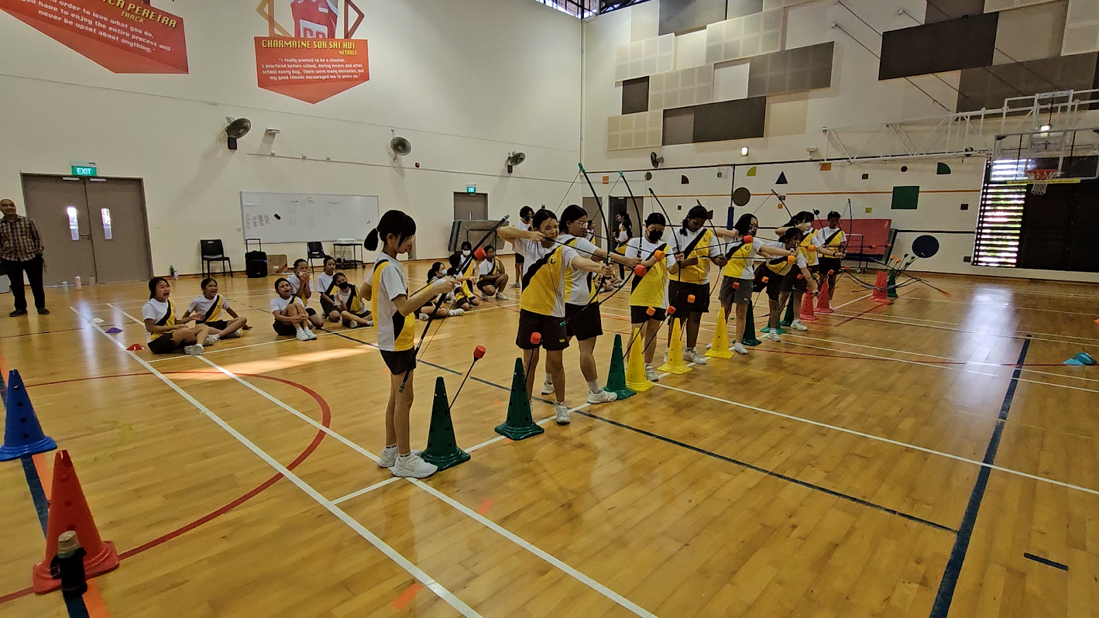 Primary school students in yellow and white PE attire practising archery indoors in a school hall. A row of students stands at green traffic cones, drawing bows with foam-tipped arrows, while another group sits on the floor watching. Motivational banners featuring alumni quotes are displayed on the wall behind them.