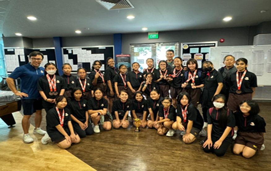 A large group of students in black uniforms wearing medals pose indoors with a small trophy on the floor.