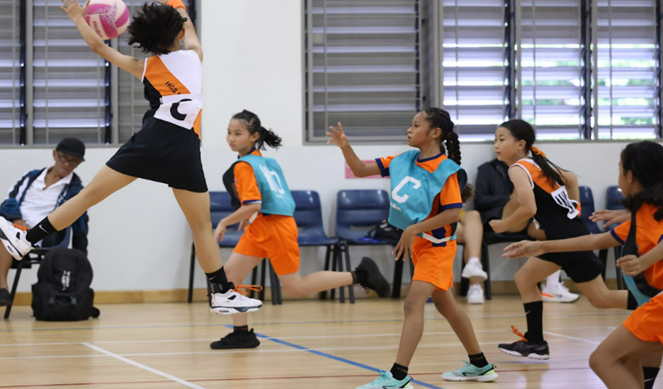 Young girls playing indoor netball, one jumping to throw a pink ball while others defend on a wooden court.