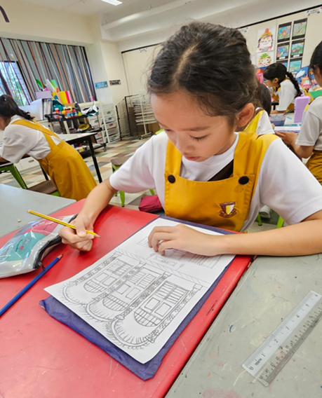 Young girl in yellow apron draws a building sketch with pencil at a red desk in a colorful classroom.