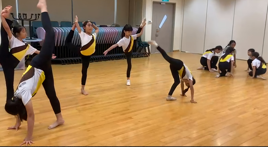 Girls in matching white, black, and yellow uniforms perform gymnastics and dance moves on a wooden floor indoors.