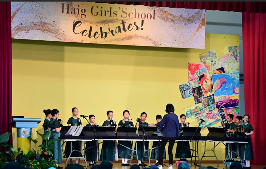 Students in green uniforms perform on stage at Haig Girls School Celebrates event, led by a conductor.