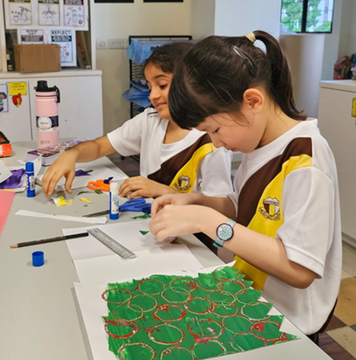 Two young girls in school uniforms work on an art project at a table with glue sticks, a ruler, and colored paper.