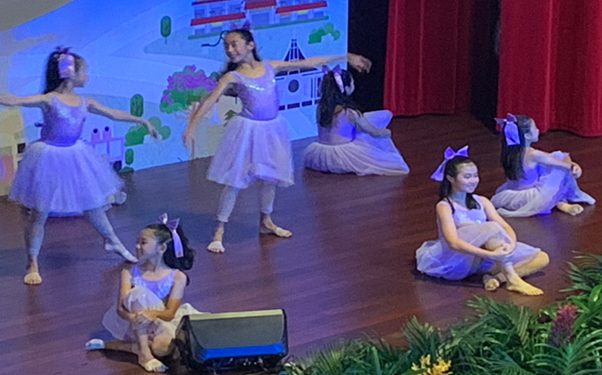 Young girls in purple tutus performing a dance recital on a wooden stage with a colorful backdrop.