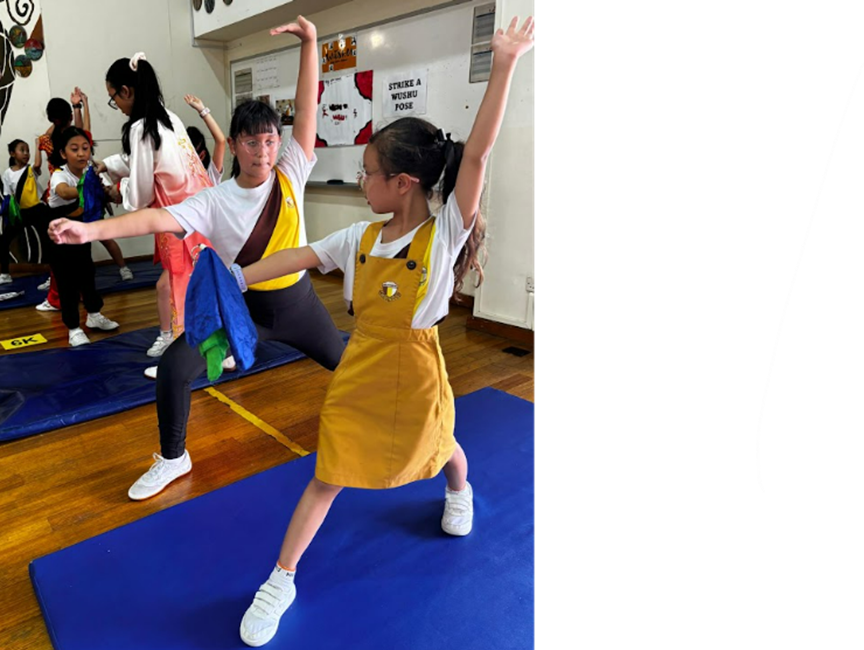 Children and an adult practice Wushu poses on blue mats in a gym with a "Strike a Wushu Pose" sign on the wall.