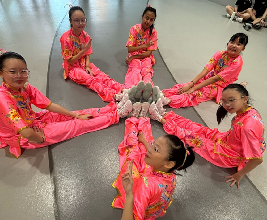 Six girls in bright pink embroidered uniforms sit in a circle on the floor with feet touching at the center.