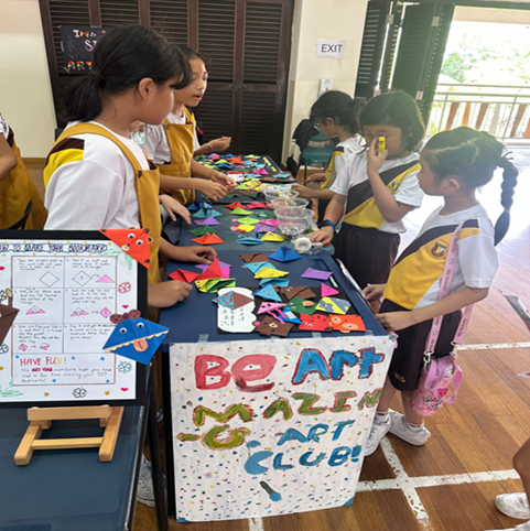 Students in school uniforms browse colorful origami and crafts on a table at an Art Club display booth.