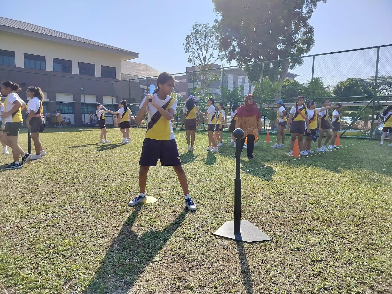 A student in school PE uniform prepares to bat a ball off a tee on a grass field, with classmates and a teacher in a red hijab visible in the background during an outdoor PE lesson.