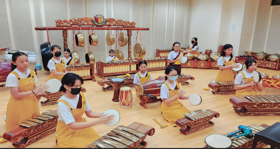 Children in yellow aprons play traditional gamelan instruments including gongs, xylophones, and hand drums indoors.