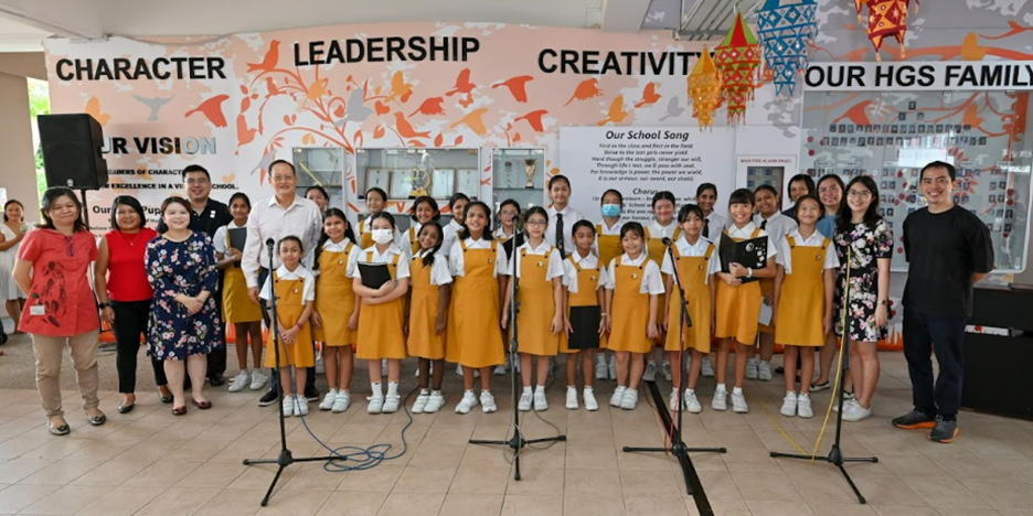 A school choir of girls in yellow uniforms stands at microphones before a banner reading Character, Leadership, Creativity.