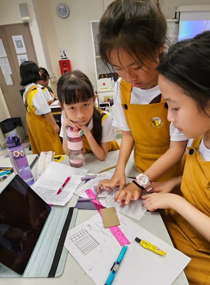 Three young girls in yellow school uniforms gather around a desk with papers, rulers, and a tablet in a classroom.
