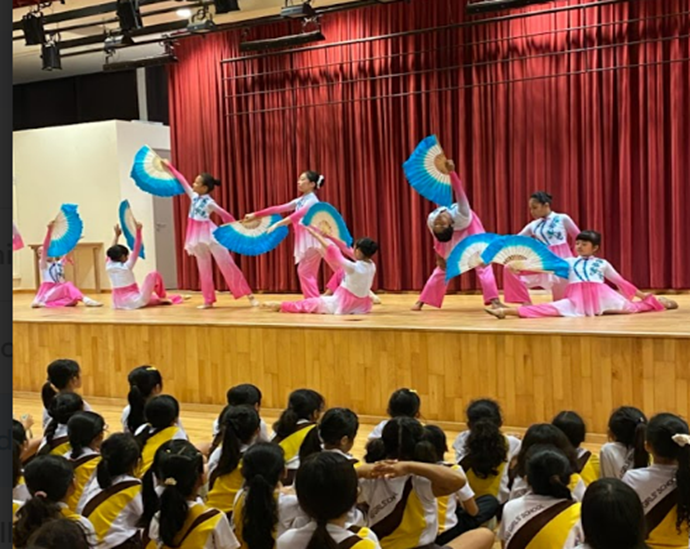 Girls in pink and white costumes perform a fan dance on stage before a seated audience of children.