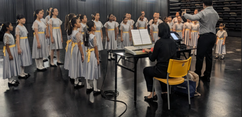Girls in gray dresses with yellow sashes stand in choir formation led by a conductor while a pianist plays.