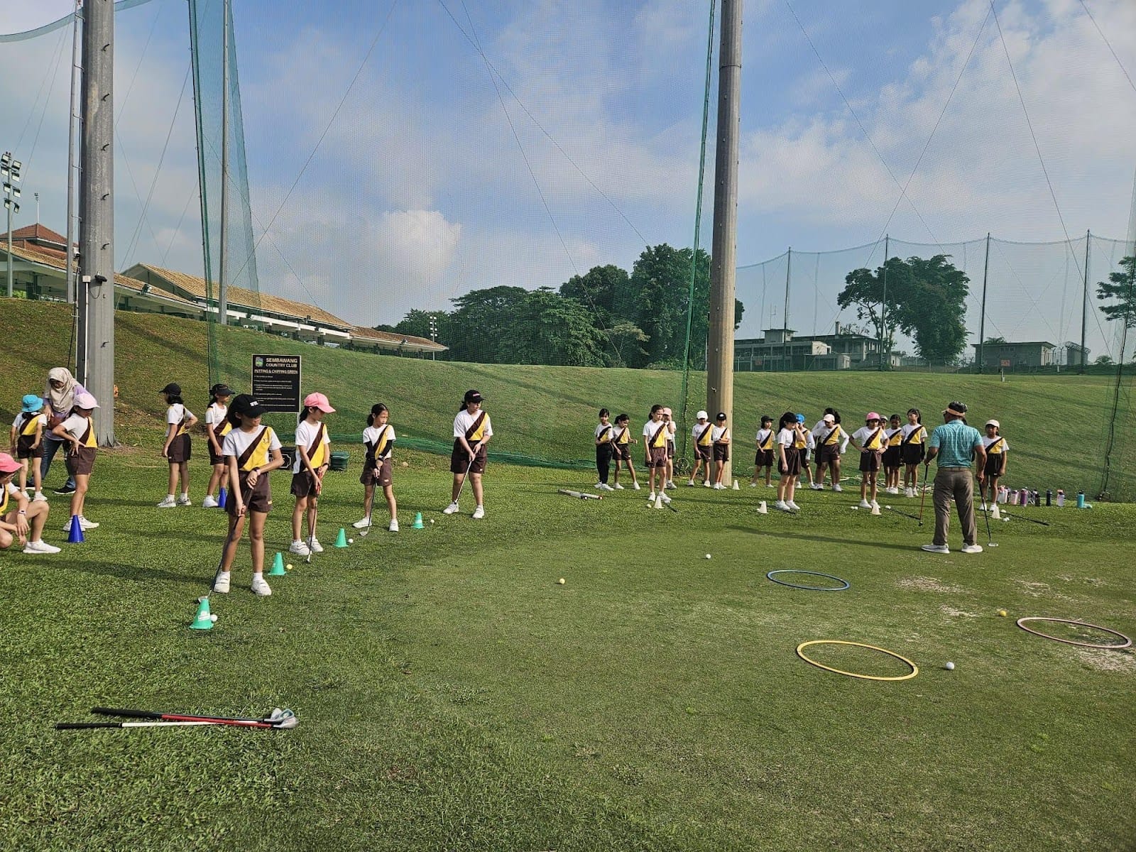 Primary school students in PE attire receiving a golf lesson on the driving range of Sembawang Country Club. Students stand in a line on the grass, holding golf clubs, while an instructor demonstrates technique. Hula hoops and cones are laid out as training aids, with tall netting surrounding the range in the background.