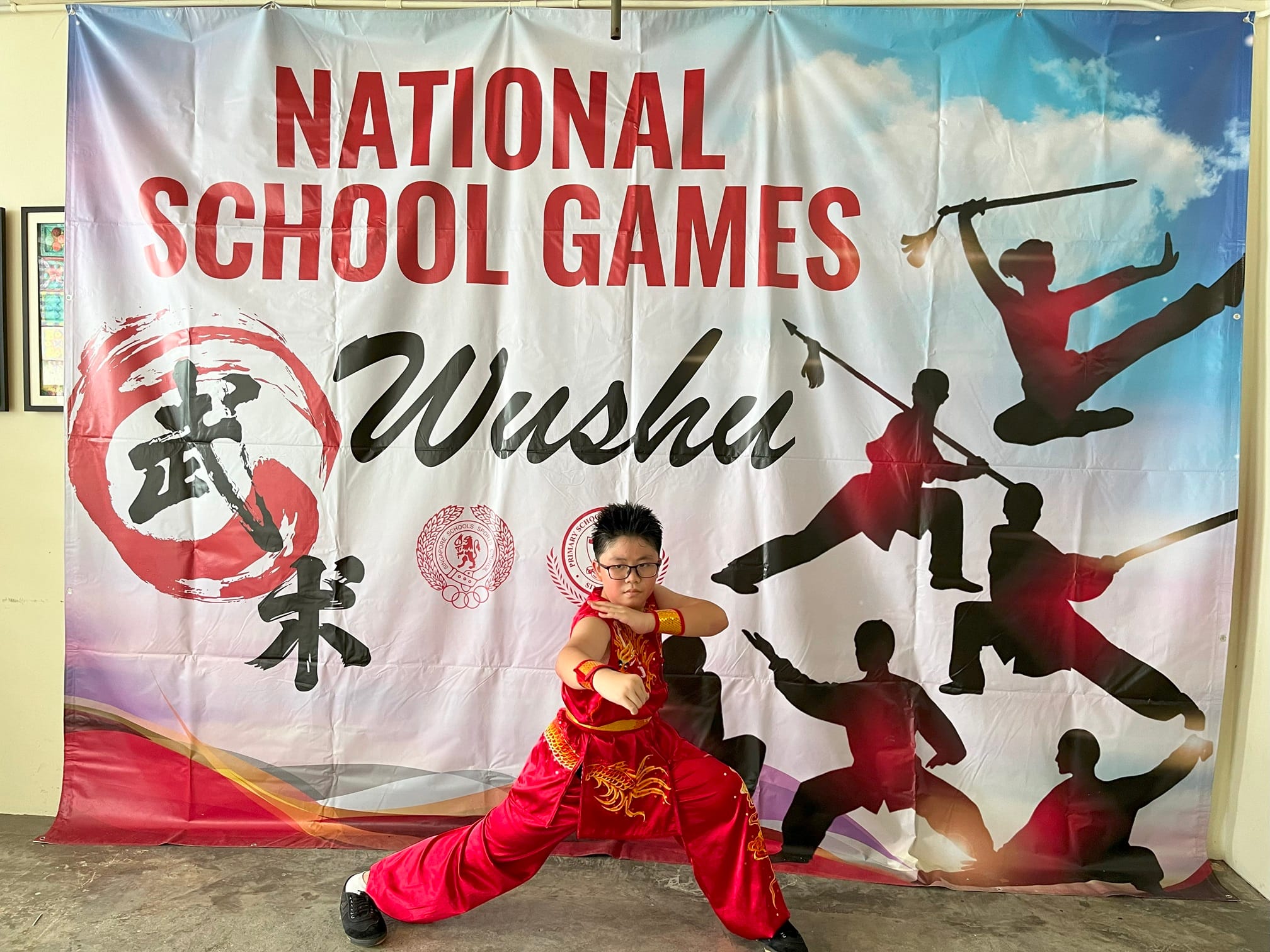 Boy in red wushu uniform poses in a martial arts stance before a National School Games Wushu banner.