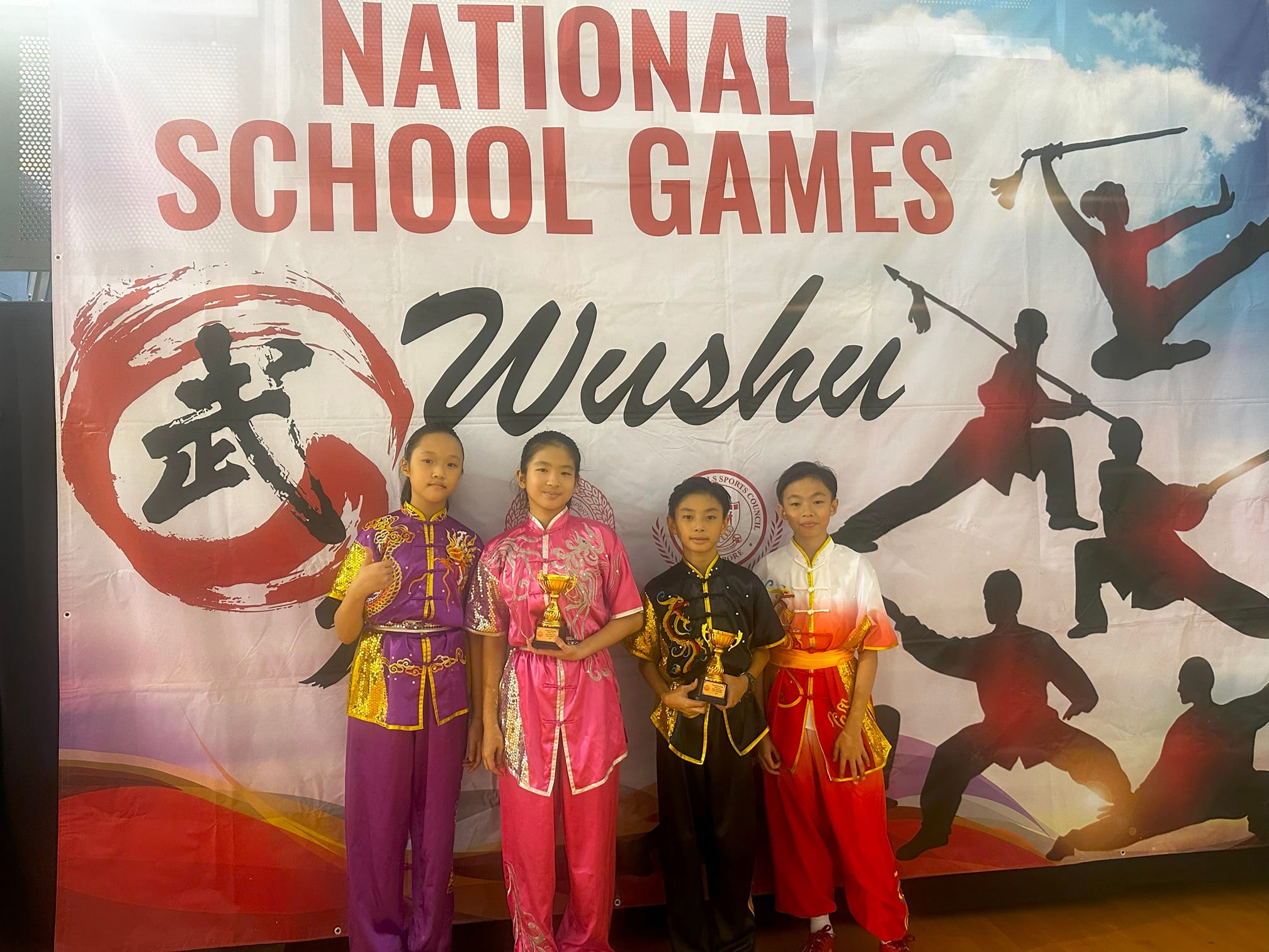 Four children in colorful wushu uniforms hold trophies in front of a National School Games Wushu banner.