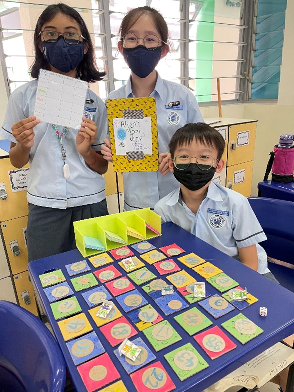 Three masked students in school uniforms display a handmade number board game with colorful numbered tiles on a blue desk.
