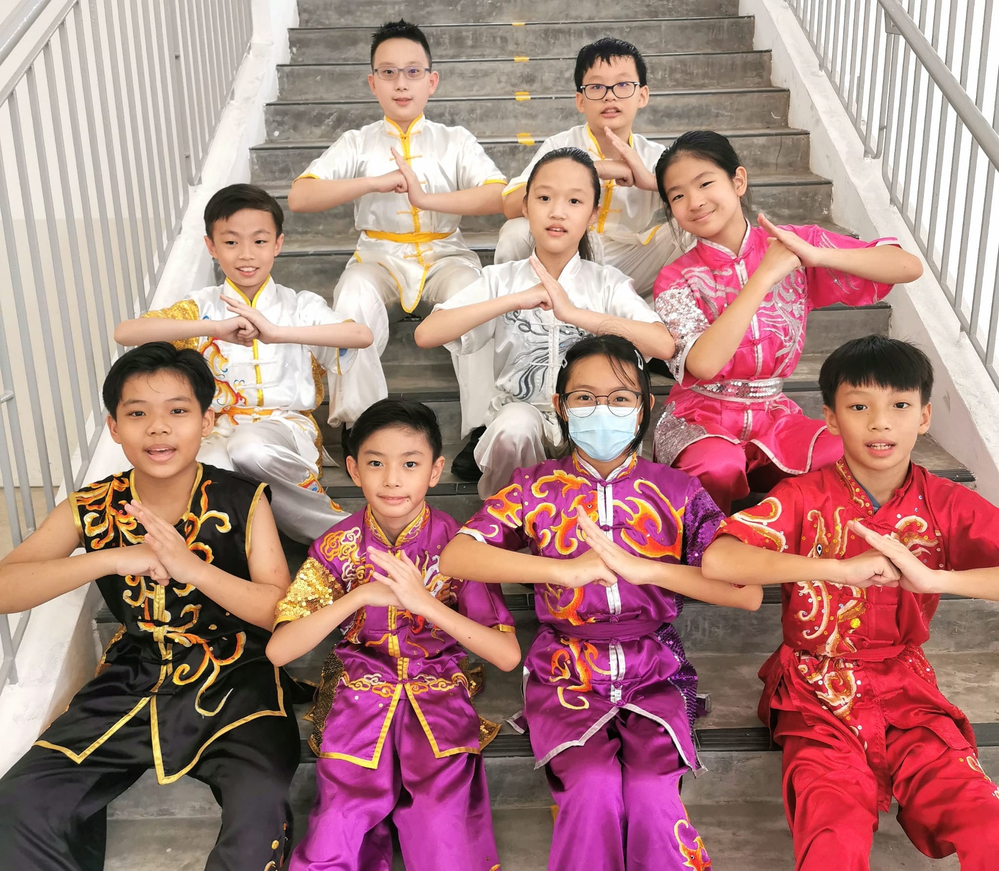 Nine children in colorful wushu uniforms pose with martial arts salute gestures on a staircase.