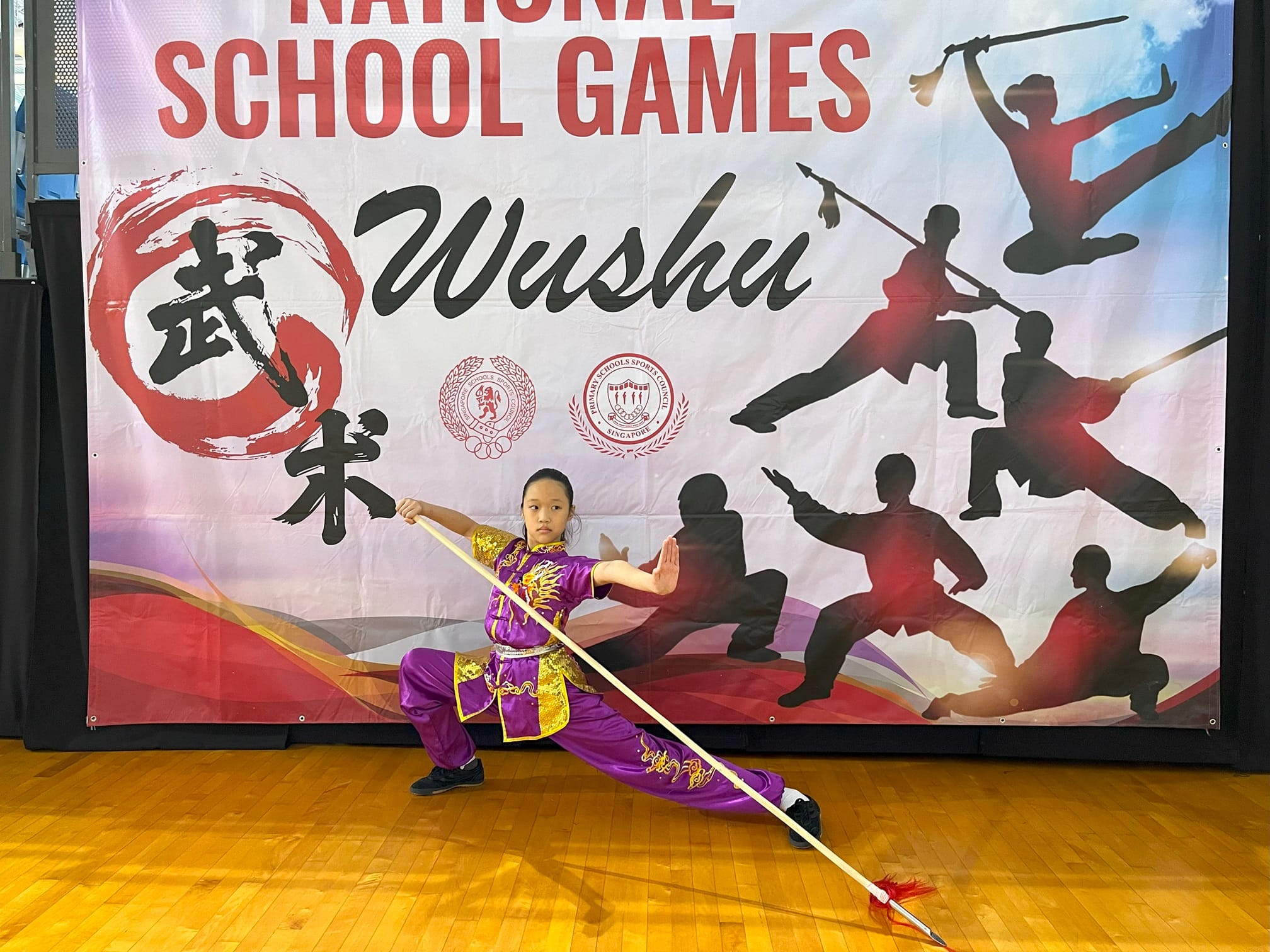 A young girl in a purple and gold wushu uniform holds a spear in a low stance before a National School Games Wushu banner.
