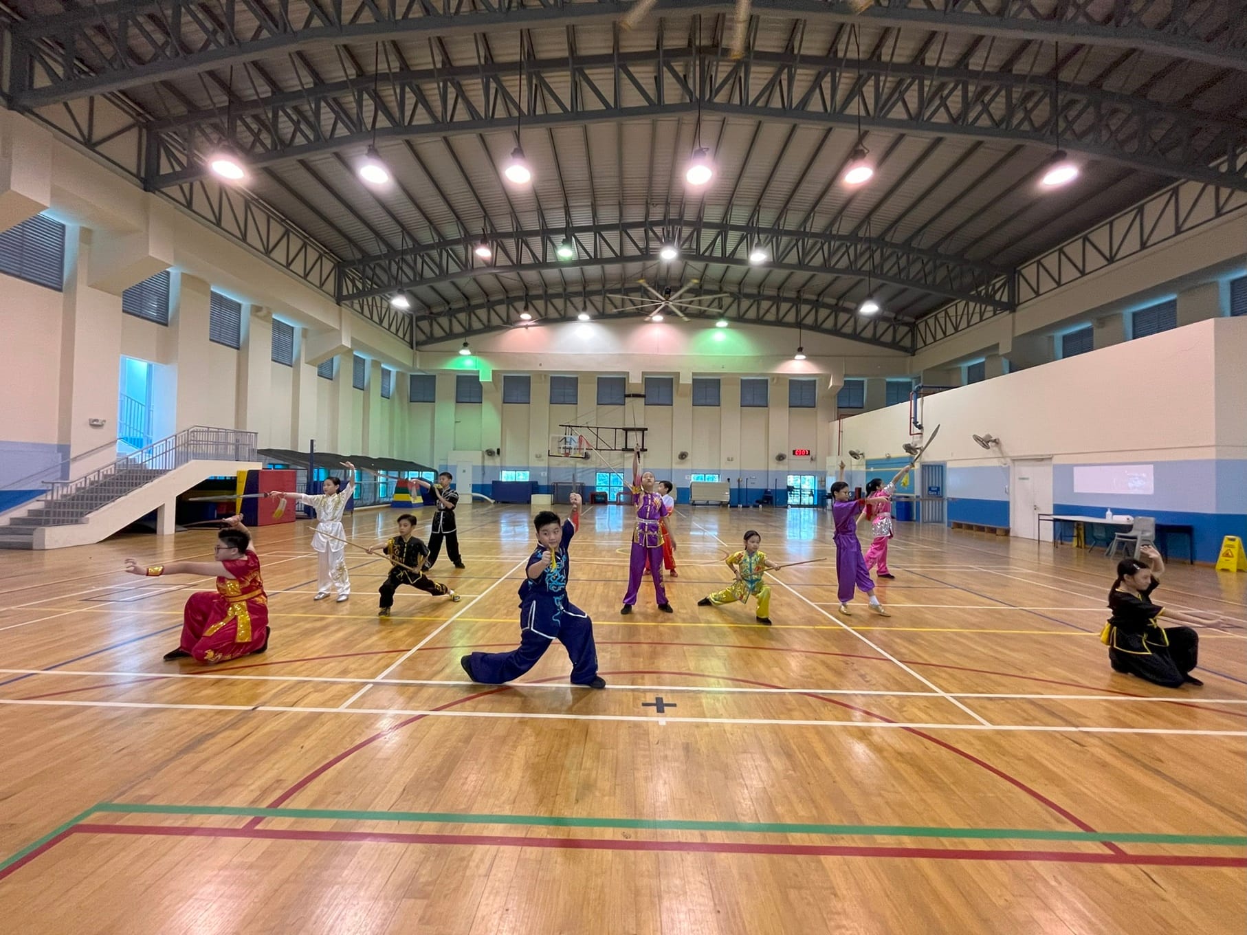 Children in colorful martial arts uniforms practice wushu with weapons on a wooden gymnasium floor.