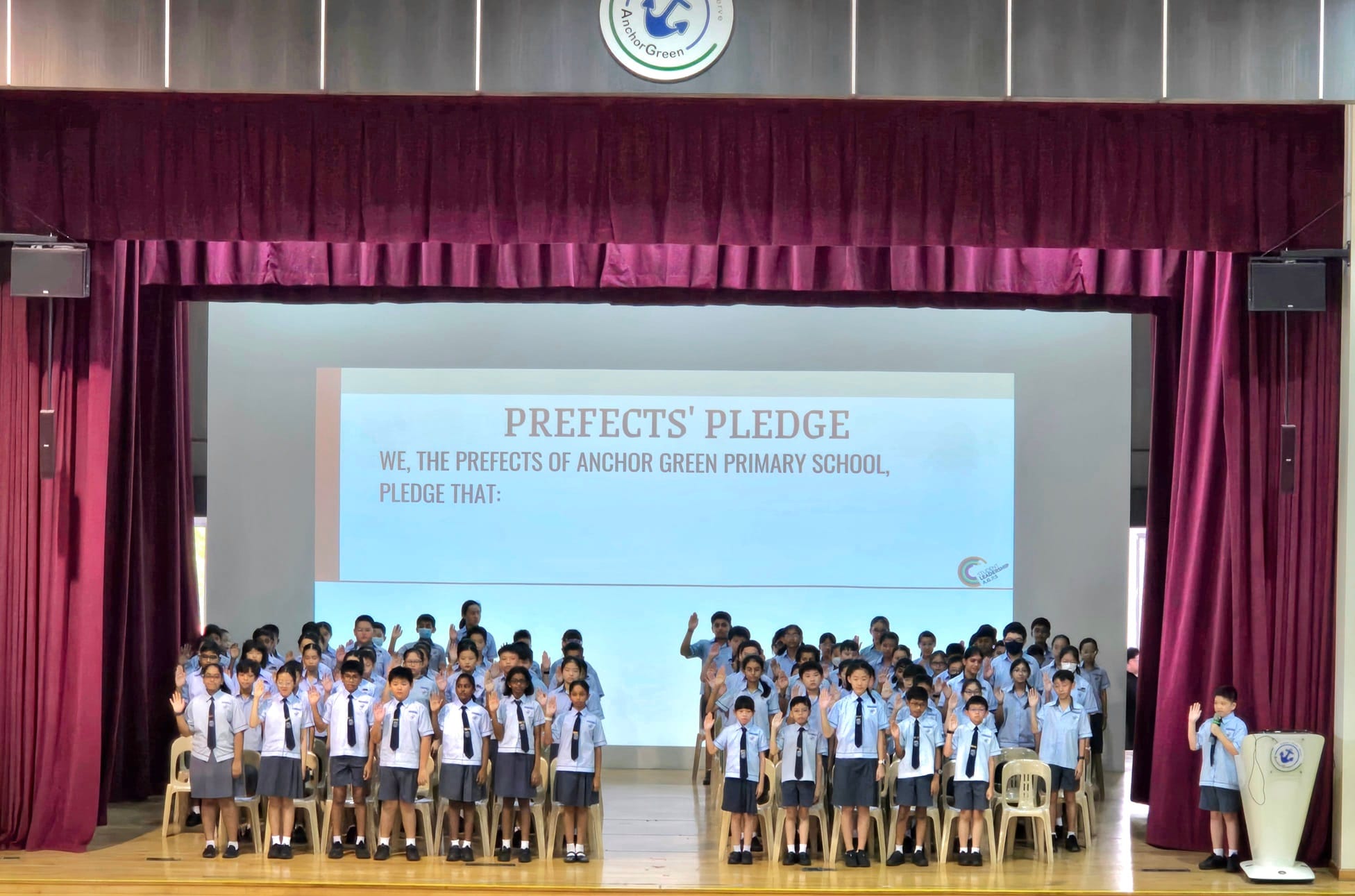 Students in uniforms raise their hands on a school stage during the Anchor Green Primary School Prefects' Pledge ceremony.