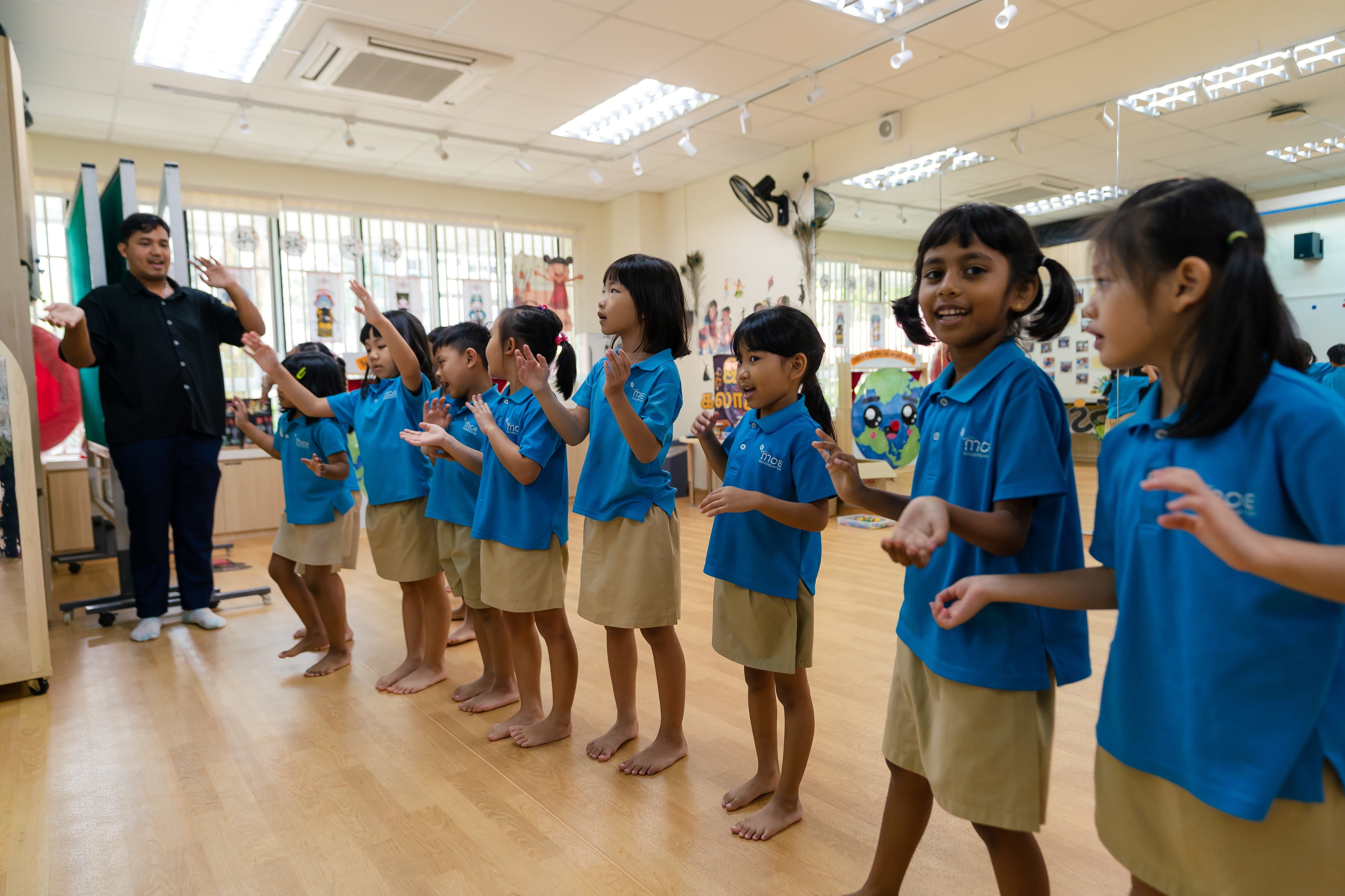 Teacher leads a line of barefoot children in blue shirts and tan shorts doing hand motions in a classroom.
