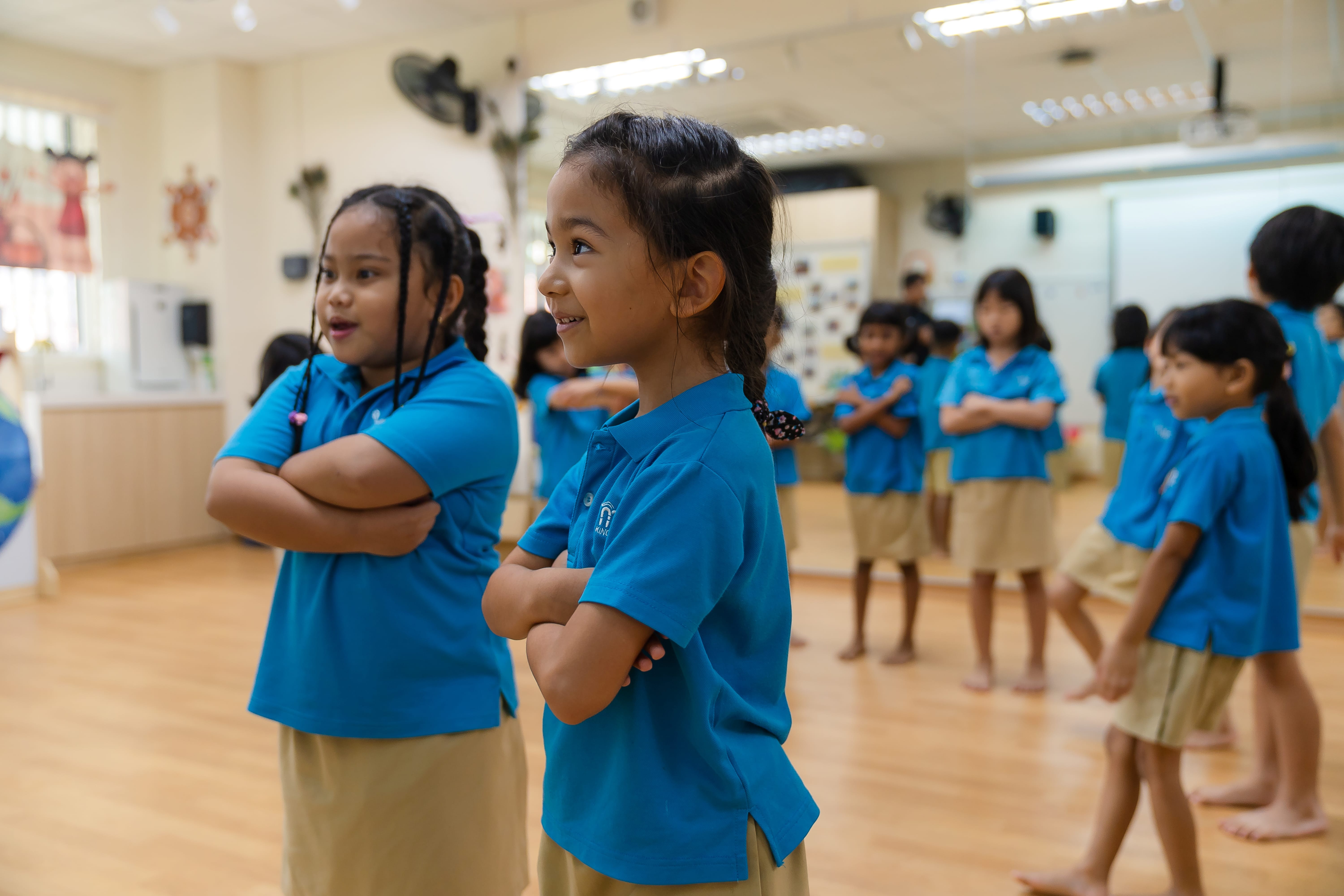 Schoolchildren in blue uniforms stand with arms crossed in a classroom, two girls smiling in front.
