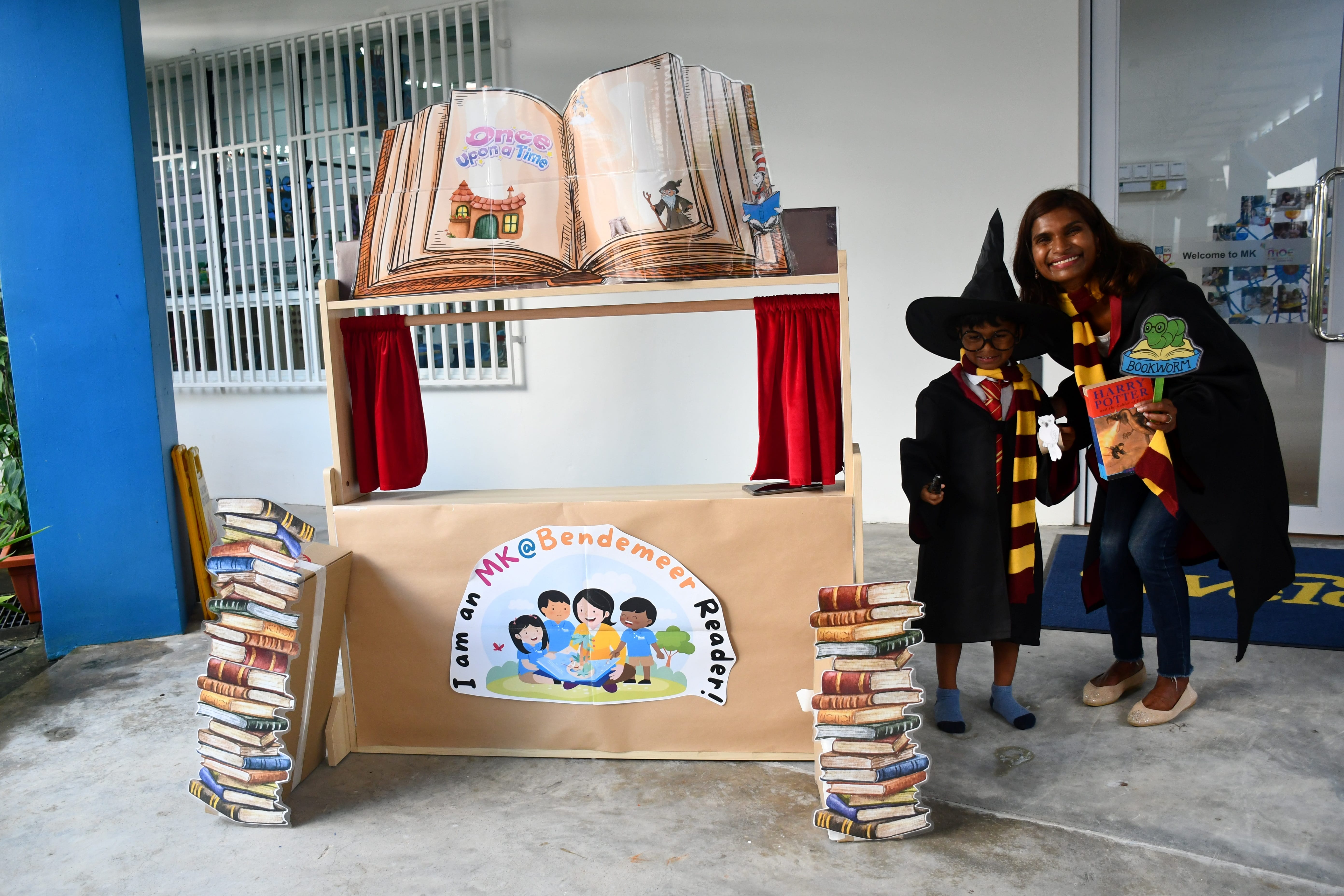 Woman and child in wizard costumes pose by a book-themed booth with stacks of books and a giant open book.