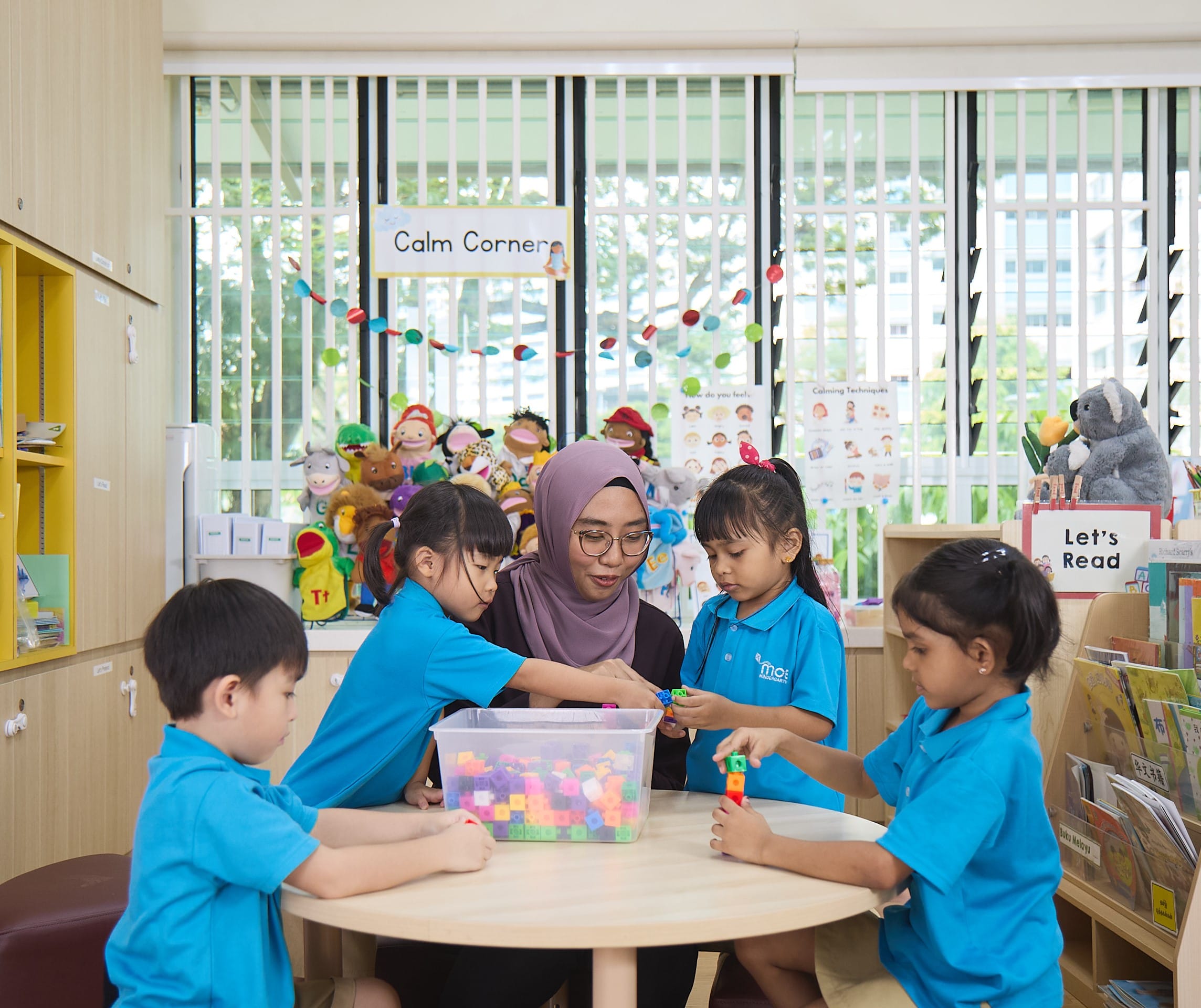 Four young children in blue uniforms playing with colorful blocks in a bright classroom.