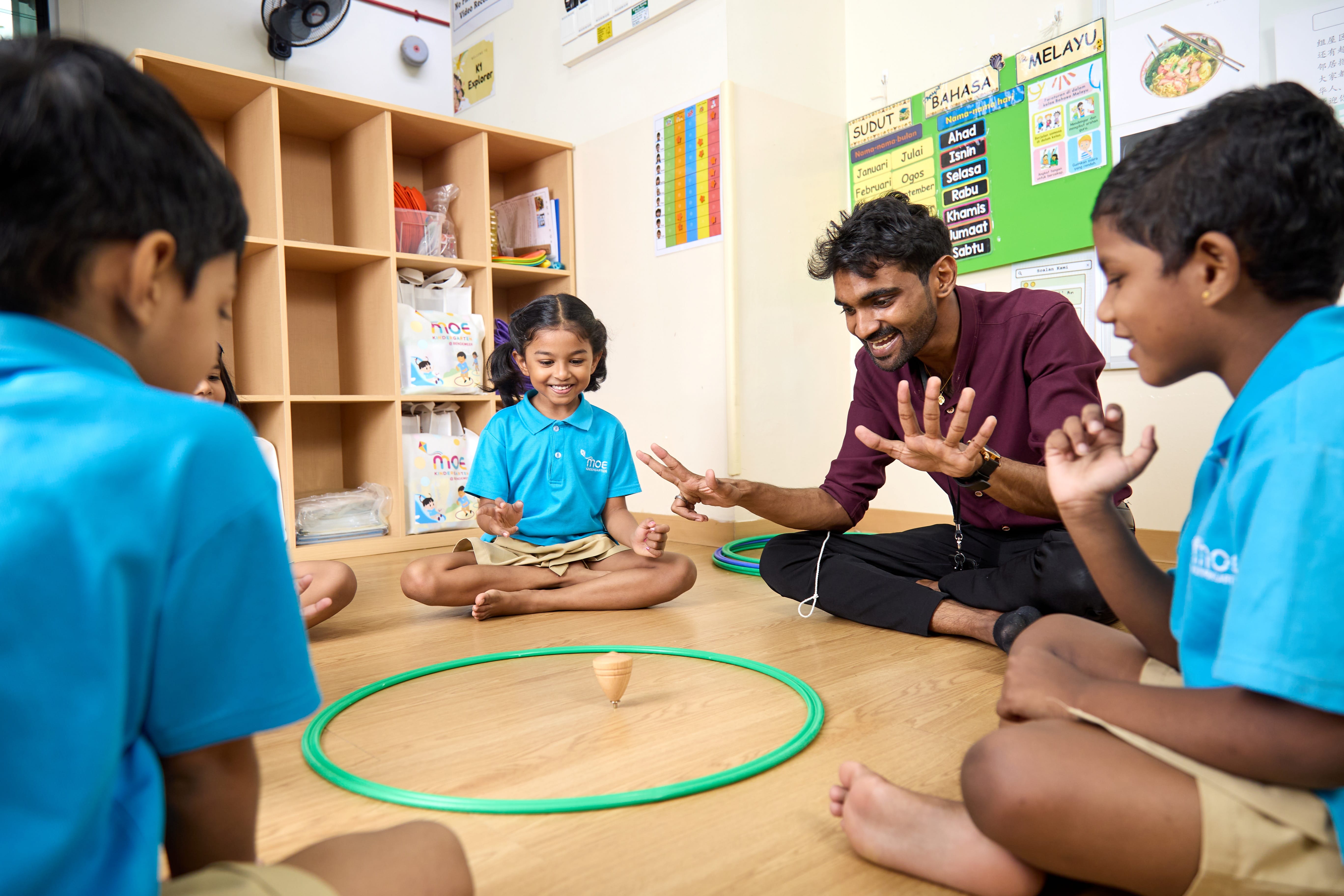 Teacher and children sit on the floor watching a spinning top inside a green hoop in a classroom.