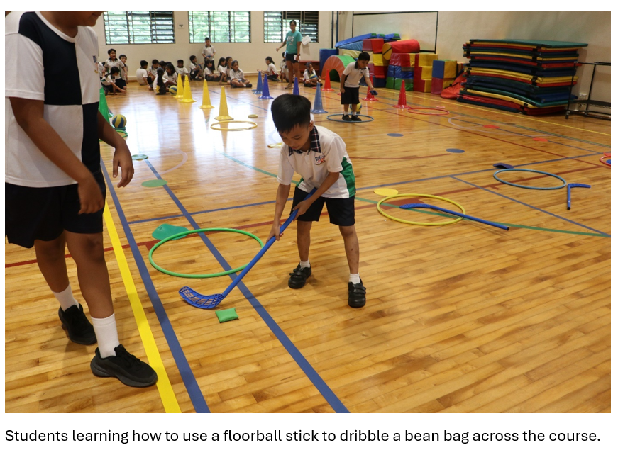 Children in a school gym use floorball sticks near hoops and cones on a wooden court.