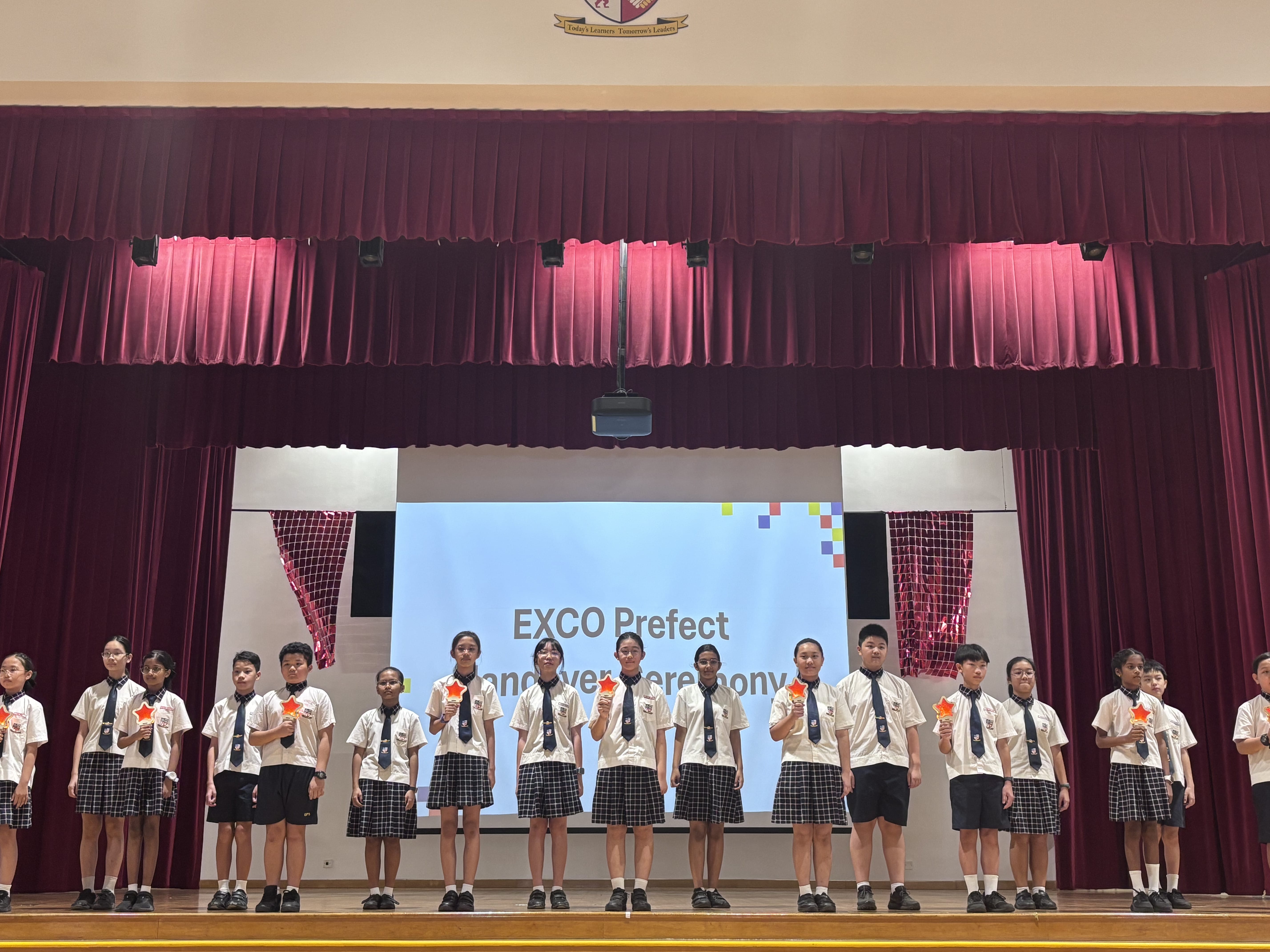 Students in school uniforms on stage before a screen reading EXCO Prefect Inauguration Ceremony.