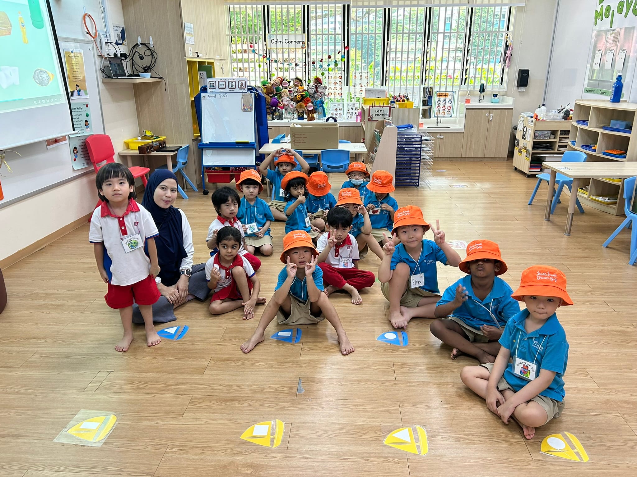 A teacher and young children in blue uniforms and orange hats sit on a classroom floor, smiling at the camera.